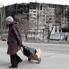 Une femme âgée marche avec un chariot devant un grand immeuble à logement endommagé par les bombes. 