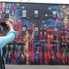 Une femme prend une photo d'une grande murale qui décore un édifice de trois étages. L'oeuvre colorée représente des gens qui marchent sous des parapluies.