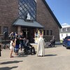 Un prêtre devant l'église, des gens en fauteuil roulant, des voitures qui défilent devant l'église.