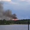 Un panache de fumée s'élève dans le ciel. Des flammes dépassent les arbres près d'un lac.