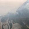 Des volutes de fumée s'élèvent d'une forêt située sur le flanc d'une montagne. Le nuage de fumée ainsi formé jette un voile épais sur une route et la voie ferrée qui longe une rivière à la base de la montagne.