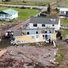 Photo aérienne de chalets et de maisons mobiles endommagées par l'ouragan. Au milieu, une maison n'a plus de toit et deux hommes sont debout devant un grand trou dans le mur.