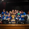 Treize personnes vêtues de chandails bleus et avec des médailles dorées au cou posent ensemble assis autour d'un trophée.