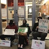 Quatre jeunes sont assis sur des chaises et tiennent des pancartes devant les portes vitrées d'un établissement.