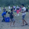 Des enfants jouent dans un espace vert et on aperçoit des adultes au fond.