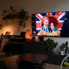 Une femme dans son salon tient une télécommande dans sa main et regarde un écran de télévision sur lequel est affiché le couple royal formé de Harry et Meghan. 
