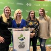 Sami Jo Small, Heather Morrison, Vicki Keith et Heather Moyse posent en souriant pour une photo entre un grand mur vert et un podium décoré du logo des Jeux du Canada 2023.