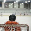 Un enfant est gardien de but de hockey. Il est de dos et fait face au reste des joueurs sur une patinoire intérieure.