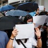 Plusieurs personnes réunies dans la rue, portant des masques, des pages blanches et des parapluies.