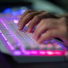 Des doigts tapent sur un clavier d'ordinateur qui est éclairé par des néons roses et mauves.
