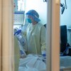 Une infirmière qui porte une jaquette jaune des gants bleus, un bonnet bleu, un masque et une visière est au chevet d'un lit d'hôpital.