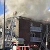 Des flammes et beaucoup de fumée sortent du toit d'un immeuble. Un pompier tente d'accéder au toit avec une échelle sur un camion.