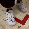 Plan rapproché des chaussures que porte une infirmière. Les chaussures sont blanches et sont faites d'un matériel dont l'imprimé est décoré de petits dessins liés à sa profession, comme des pansements, des stéthoscopes, des seringues et des thermomètres. Les mots « Mom Nurse » sont aussi écrits sur les chaussures.