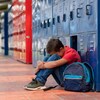 Un enfant assis par terre la tête entre les genoux et le dos à des casiers dans le corridor d'une école.