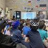Des jeunes sont assis dans un gymnase devant quelqu'un qui fait une présentation à l'aide d'un tableau.
