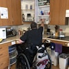Un homme en fauteuil roulant, de dos, attrape un verre dans une armoire de son appartement, alors qu'il est assis dans son fauteuil roulant.