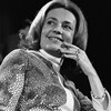 Dans un studio de télévision, Jeanne Moreau, souriante, porte un regard oblique vers sa droite, sa main gauche près de son menton.