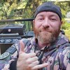 Jeremy MacKenzie, vêtu d'une combinaison de camouflage et coiffé d'une casquette à l'envers, prend un selfie à l'extérieur devant un barbecue et fait un signe de shaka avec son pouce et son auriculaire.