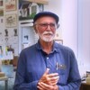 Joe Fafard se tient devant la caméra et porte un chapeau avec une chemise bleue. On voit derrière lui de nombreuses oeuvres d'art.