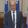 Le député pose devant un micro à l'Assemblée nationale aves des drapeaux du Québec derrière lui.