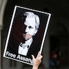 Une personne tient une affiche sur laquelle se trouve le portrait de Julian Assange muselé par un drapeau américain.