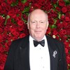 Julian Fellowes vêtu d'un complet noir posant devant un mur de roses rouges.