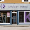 Extérieur du bureau. La lettre K est inscrite en grosse lettre dans une vitrine. Sur un panneau publicitaire au-dessus de la porte d'entrée, il est écrit : Kaméléart Matane.