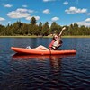 Une femme est assise dans un kayak rouge flottant sur un lac. Elle porte des lunettes fumées et des Crocs. Elle lève les deux bras dans les airs et regarde vers la rive, posant pour une photo.