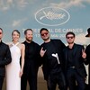 Cinq hommes vêtus de noir et une femme portant une robe blanche posent côte à côte pour les caméras sur le tapis rouge du Festival de Cannes.