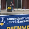 Un individu marche sur le campus de l'Université Laurentienne près d'un bâtiment, avec la pancarte de l'université en avant plan.