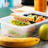 Un sandwich dans un plat de plastique, une banane et une pomme sur une table.