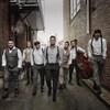 Sept musiciens habillés en habits des années 1920 déambulent dans une ruelle entre deux murs de briques.