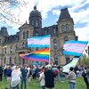 Des gens rassemblés sur la pelouse devant l'édifice de l'Assemblée législative. Des personnes agitent des drapeaux arc-en-ciel, transgenres et agenre.