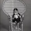 Une fillette portant des tresses, assise sur une grande chaise en paille, tient un toutou dans ses mains.