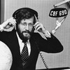 Dans un studio de radio, l'animateur Louis Martin ajuste un casque d'écoute sur sa tête. Un micro, portant une affichette avec la mention CBF 690, est suspendu devant lui.