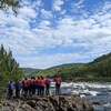 Un groupe de jeunes de dos, vêtus de gilets de sauvetage avec des pagaies à la main, sont debout face à la rivière.