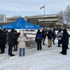 Un vingtaine de personnes prennent un café sous un chapiteau bleu en hiver. Un drapeau canadien flotte devant un gros bâtiment devant eux.