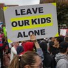 Une main tenant une affiche sur laquelle il est écrit: "Laissez nos enfants tranquilles". 