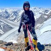 Une alpiniste attachée par des harnais regarde la caméra. On voit derrière elle des sommets enneigés.