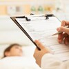 Un médecin écrit des notes dans le dossier d'un patient qu'on voit dans un lit d'hôpital en arrière-plan.