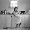 Photo en noir et blanc prise dans les années 1970 d'une jeune femme en jaquette, pieds nus, accoudée à un comptoir de cuisine couvert d'objets comme une boîte de céréales, une boîte de savon SOS et une radio. La femme étire la paupière inférieure de son oeil gauche avec son index.