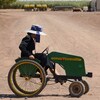 Une femme portant une robe traditionnelle mennonite et un chapeau gris au ruban bleu pédale sur un quadricycle vert de marque John Deere, sur une route de terre et de sable.