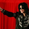 Un homme portant des lunettes fumées et des cheveux longs noirs tend son bras sur une scène ornée d'un rideau rouge, en faisant le signe de la paix avec ses doigts. 