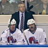 Un entraîneur est debout derrière un banc où sont assis trois joueurs de hockey. 