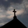 Des nuages noirs se glissent à l'arrière-plan d'une croix sur le toit d'une église catholique.