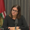 La première ministre Stefanson est assise devant deux drapeaux du Manitoba, vêtue de noir.