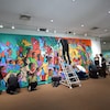 Un groupe de personnes, les unes debout, les autres à genoux, se tient face à une grande œuvre murale.