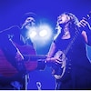 Un homme et une femme tiennent chacun une guitare dans une salle de spectacle en lumière.
