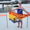Nadine Bennett en maillot de bain et pieds nus sur la glace pose pour la photo avec un drapeau du Nouveau-Brunswick dans les mains. Derrière elle, un trou a été fait dans la glace pour y faire une ligne de nage. 