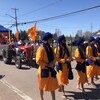 Cinq hommes habillés en jaune, pieds nus, marchent en rang en tenant un sabre. Un char allégorique tiré par un tracteur rouge suit derrière.